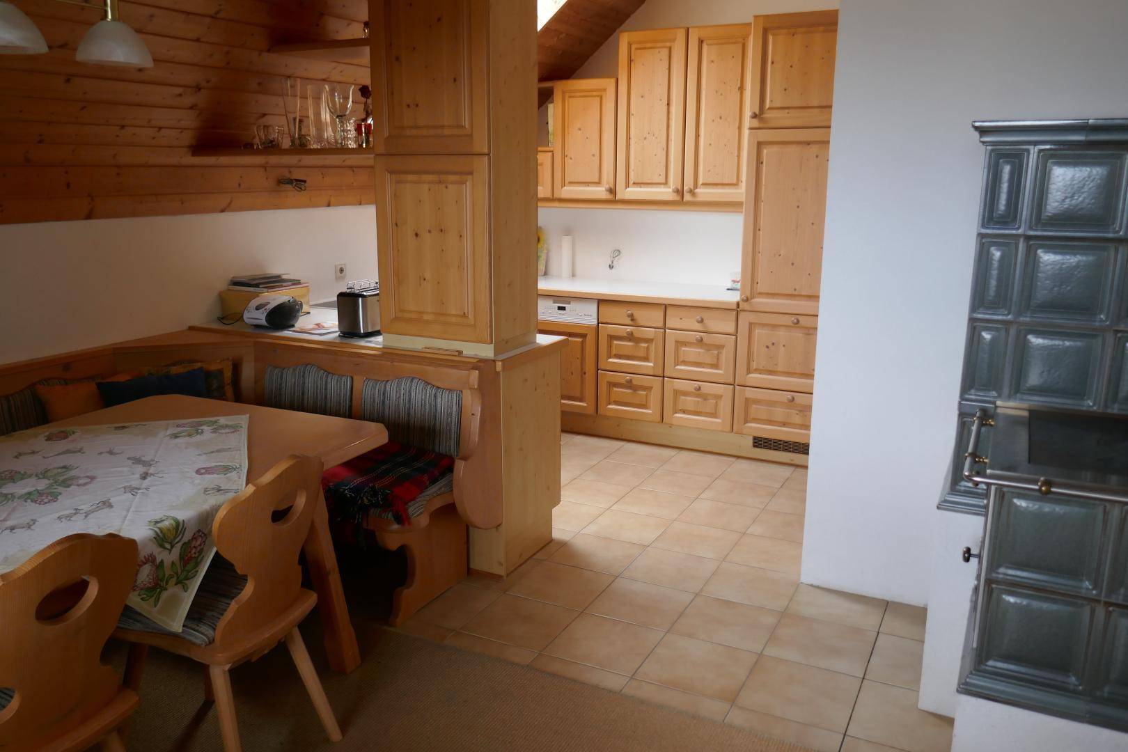Gemütliche Dachgeschossküche mit Holzbalken, Essecke und einem traditionellen Kachelofen, ideal für rustikales Wohnen.