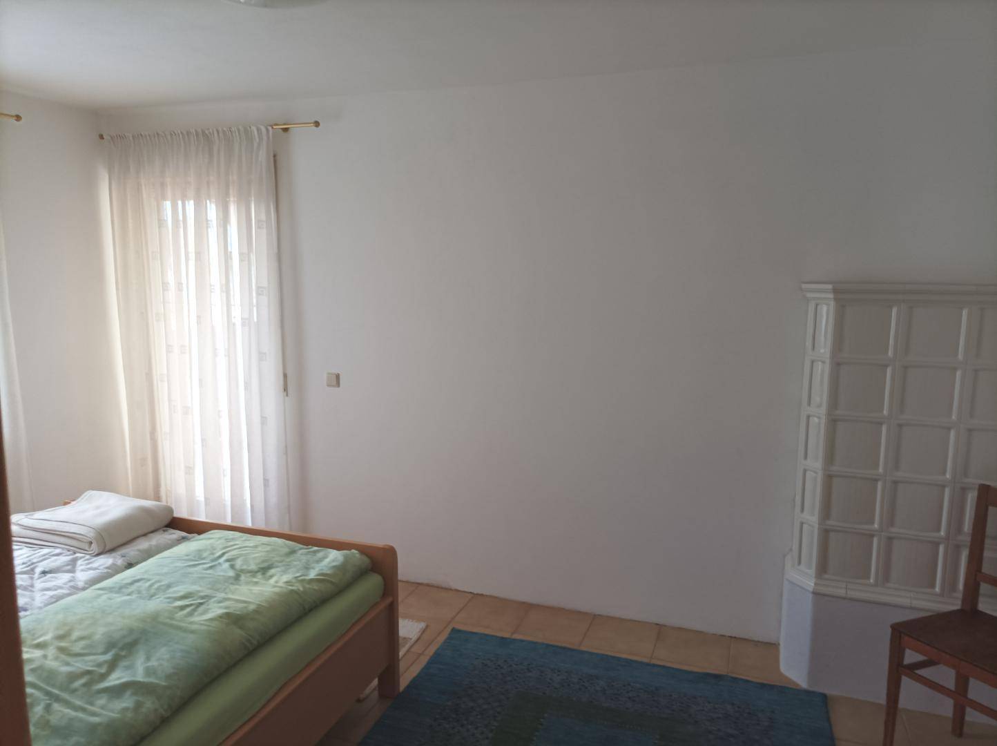 Helles Schlafzimmer mit Einzelbett, Fliesenboden und einem dekorativen Kachelofen, der Wärme spendet.