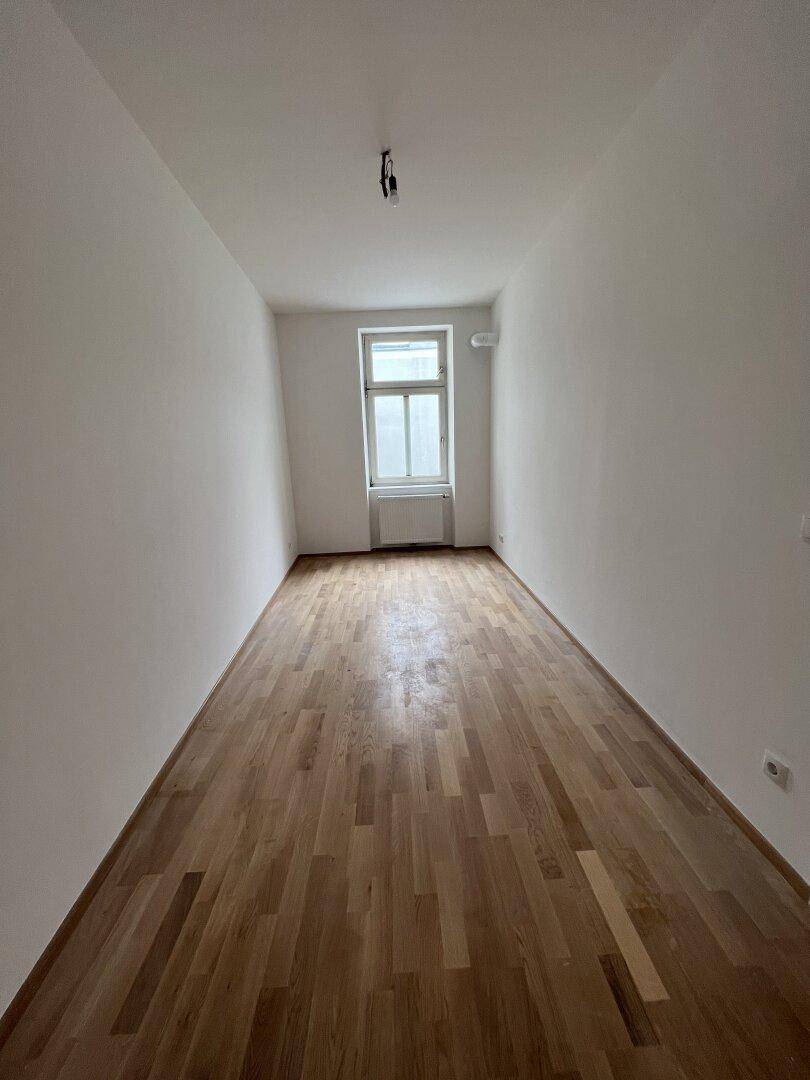 Schmaler, heller Raum mit Holzboden und einem Fenster, geeignet als Schlaf- oder Arbeitszimmer.