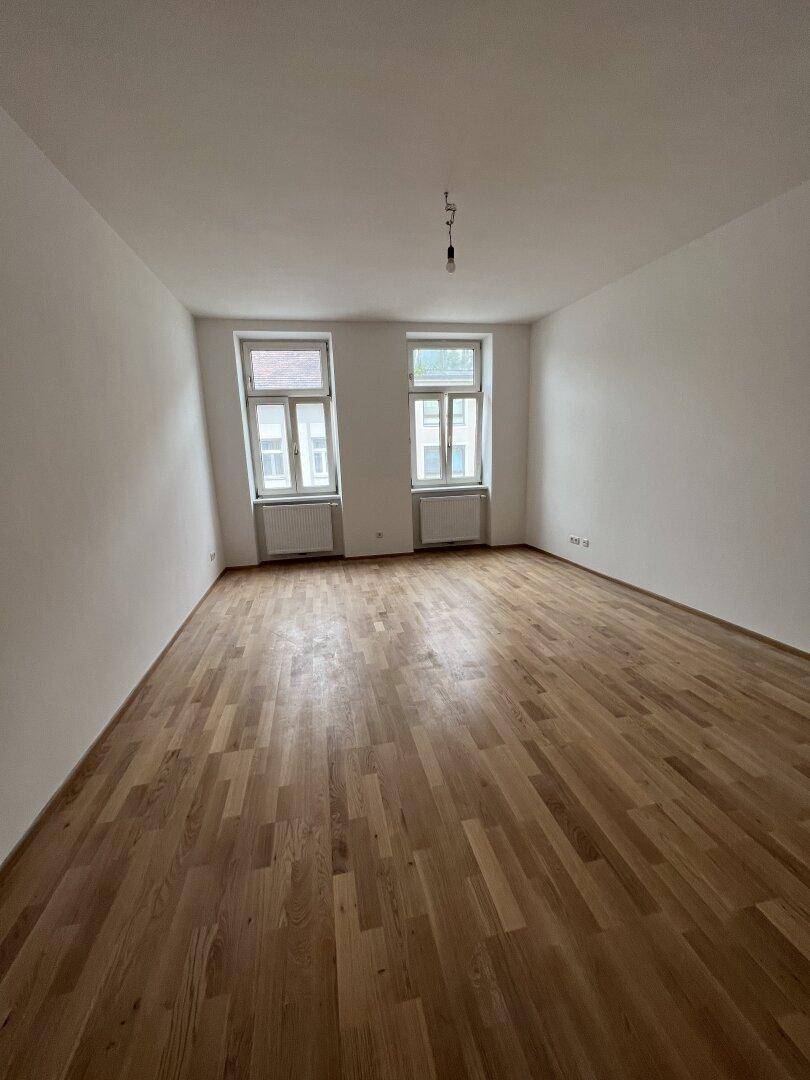 Heller, leerer Raum mit Holzboden und zwei Fenstern, bereit zur individuellen Gestaltung.