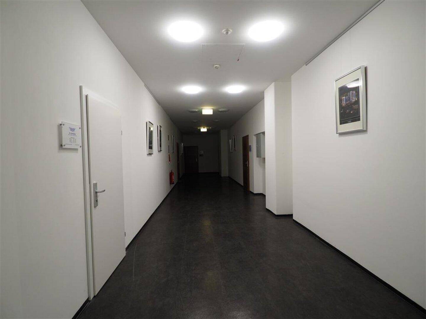 Langer, heller Flur mit weißen Wänden, dunklem Boden und mehreren Türen zu den einzelnen Räumen.