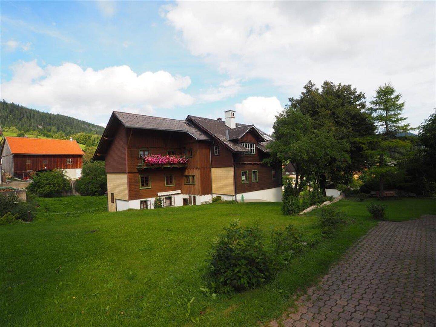 Weitläufiger grüner Garten mit traditionellem Haus und Blick auf die bewaldeten Hügel.