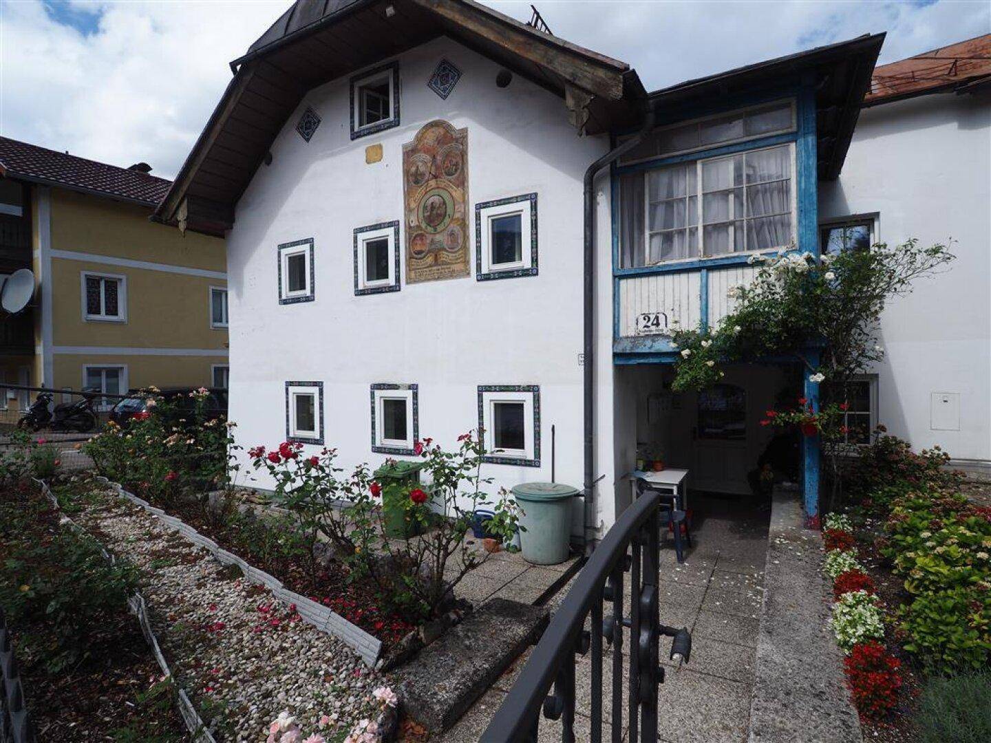 Rückseite eines traditionellen Hauses mit überdachter Terrasse und einem blühenden Rosengarten.