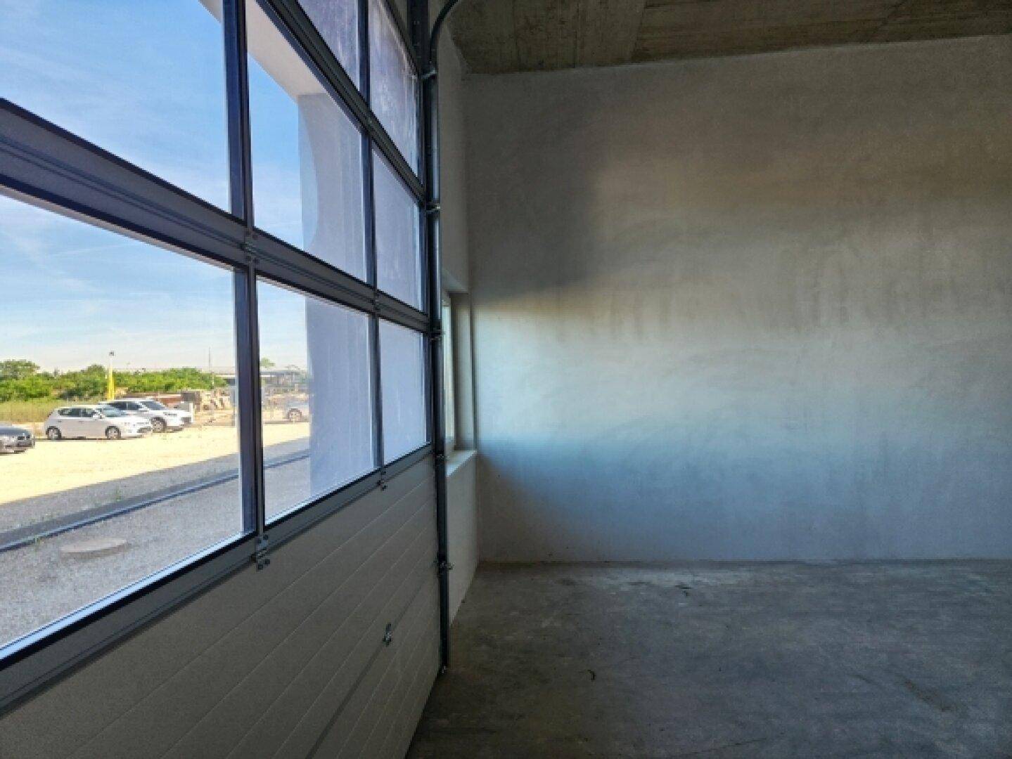 Heller Innenraum einer Halle mit großem Fenster und Blick auf den Außenbereich und parkende Autos.
