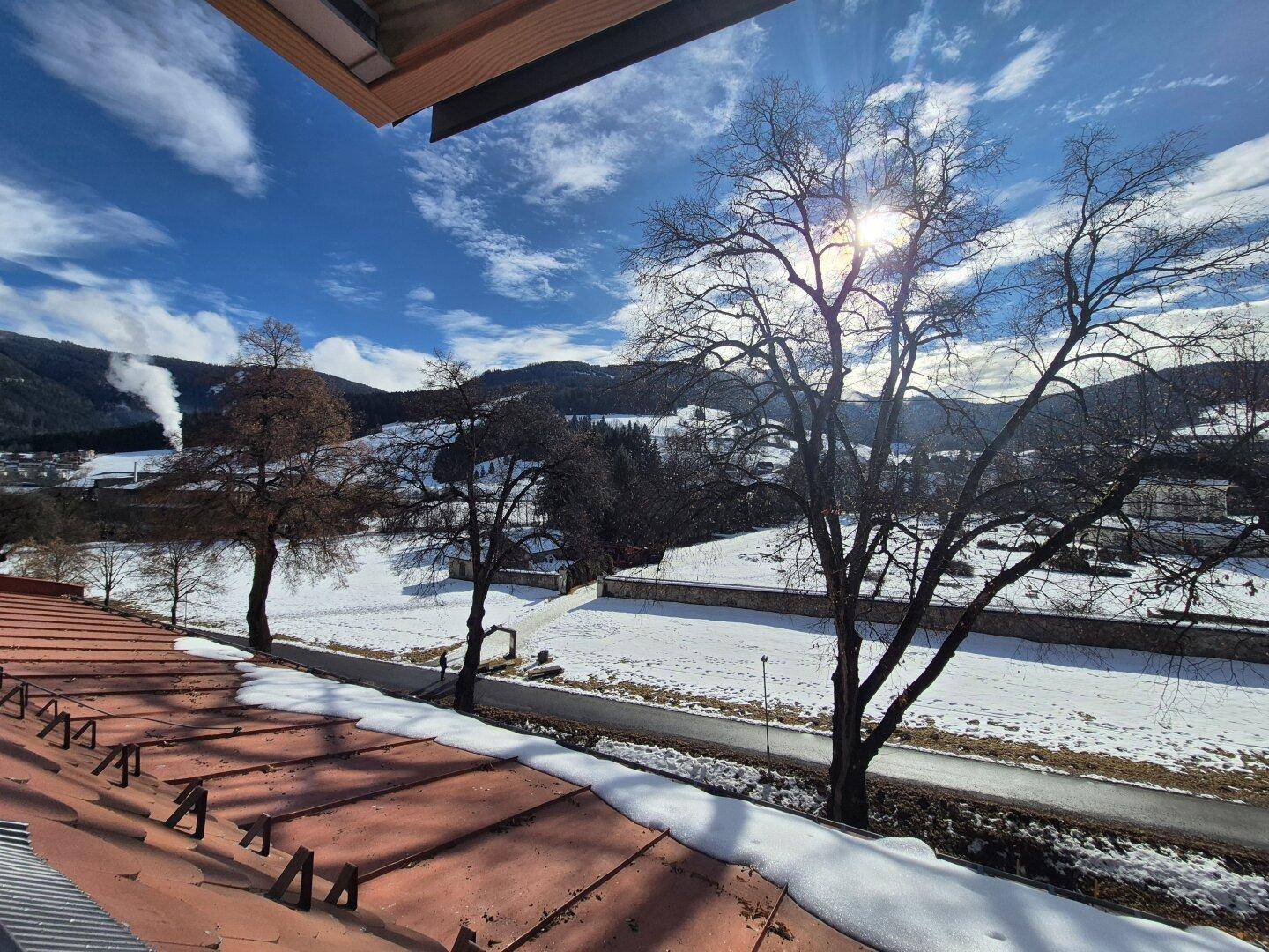 Winterliche Aussicht auf verschneite Felder und Berge von einem Dachfenster.