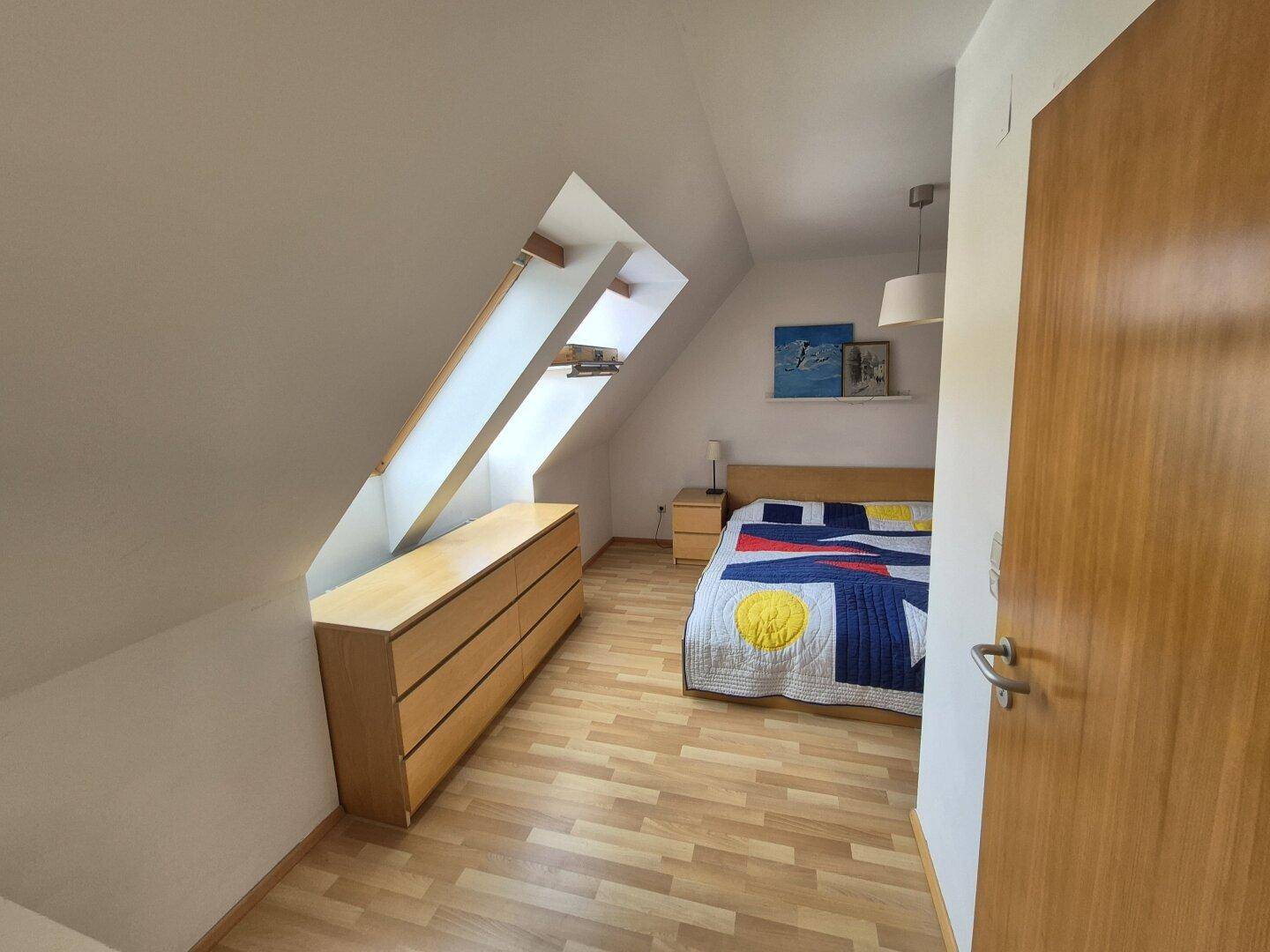 Schlafzimmer mit Holzboden, Kommode und Dachschräge, funktional eingerichtet.