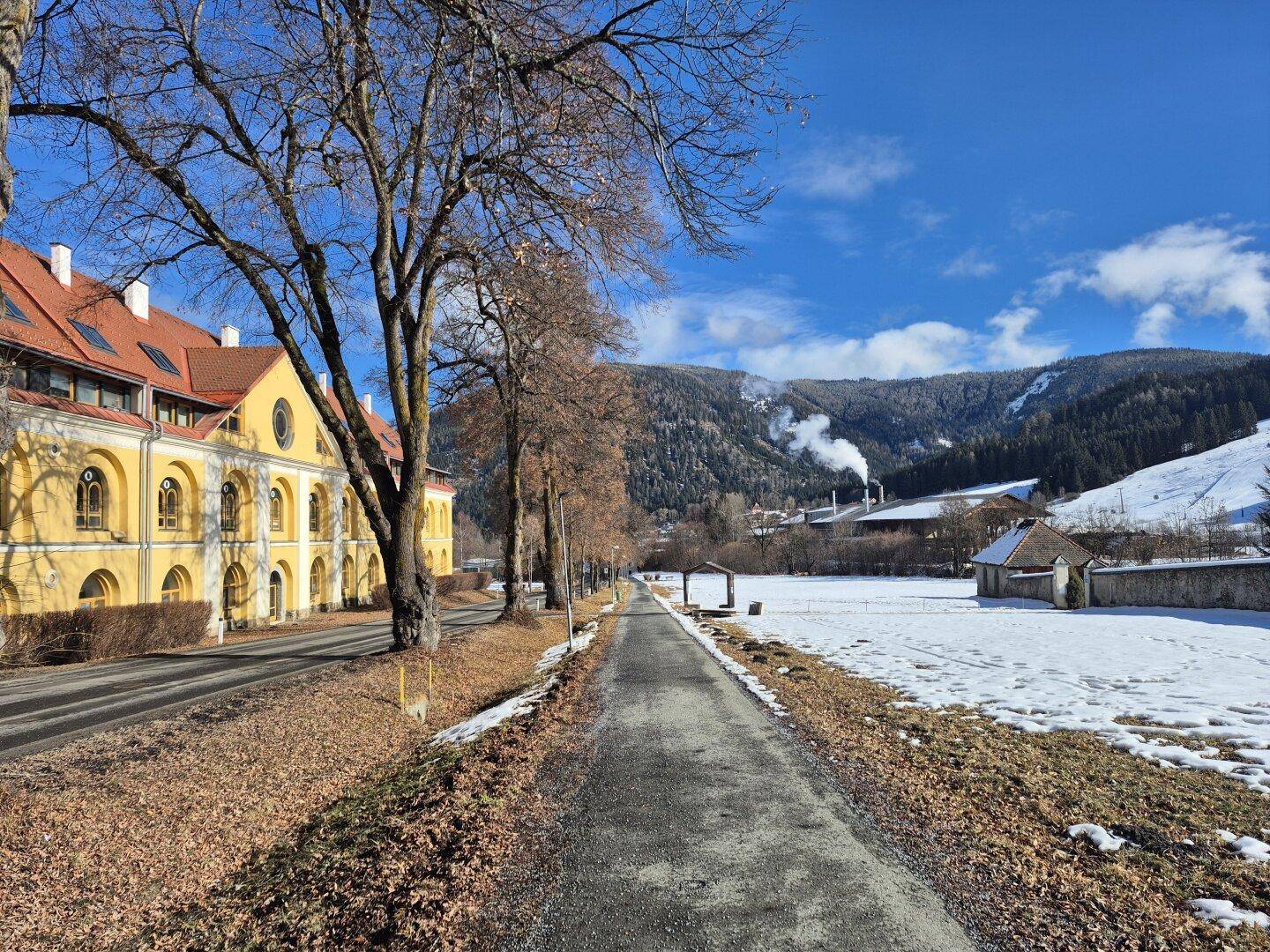 Ländliche Umgebung mit Straße, verschneiten Feldern und Bergen im Hintergrund unter blauem Himmel.