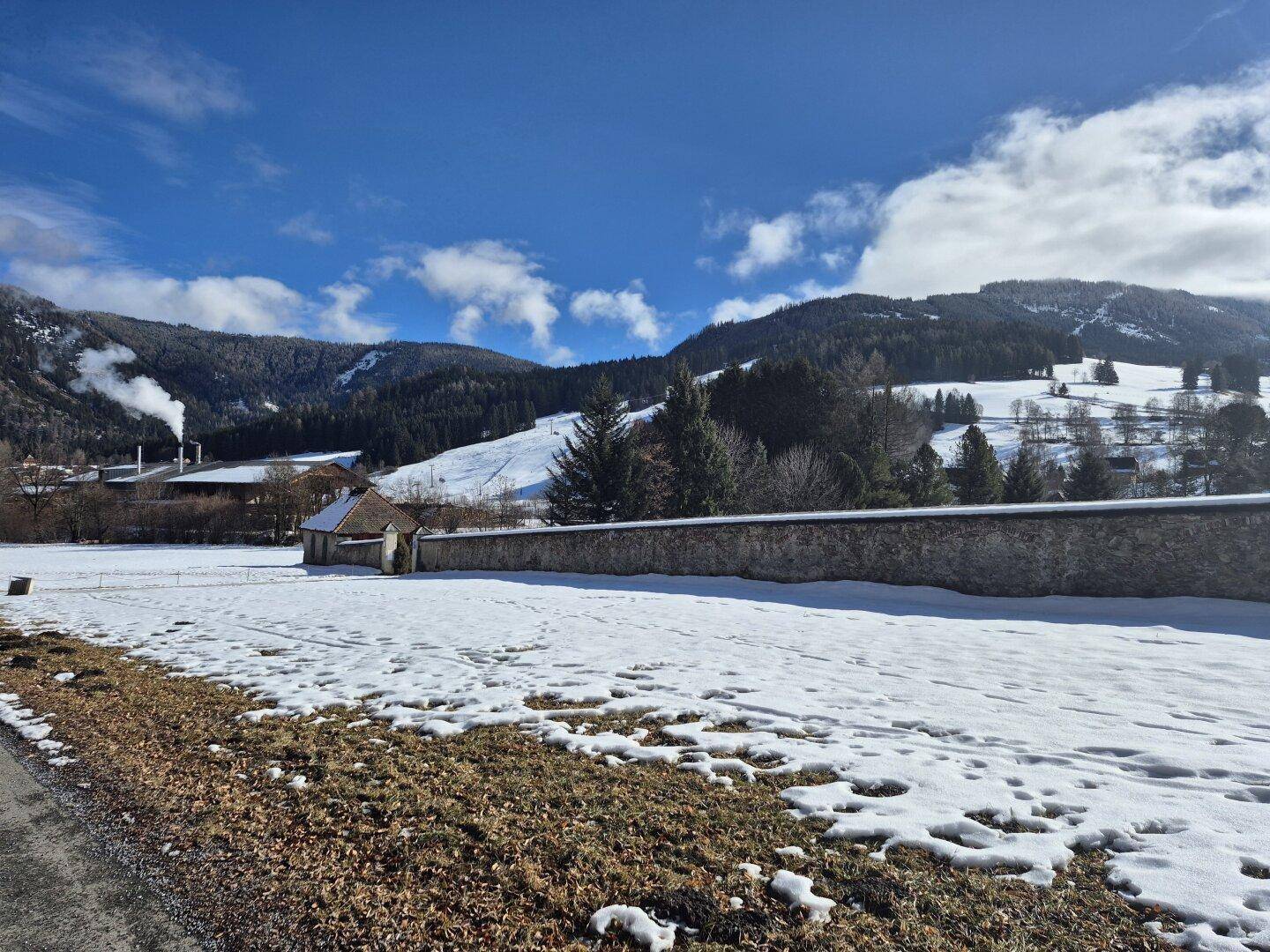 Weite, verschneite Landschaft mit Feldern, Bäumen und Bergen unter blauem Himmel.