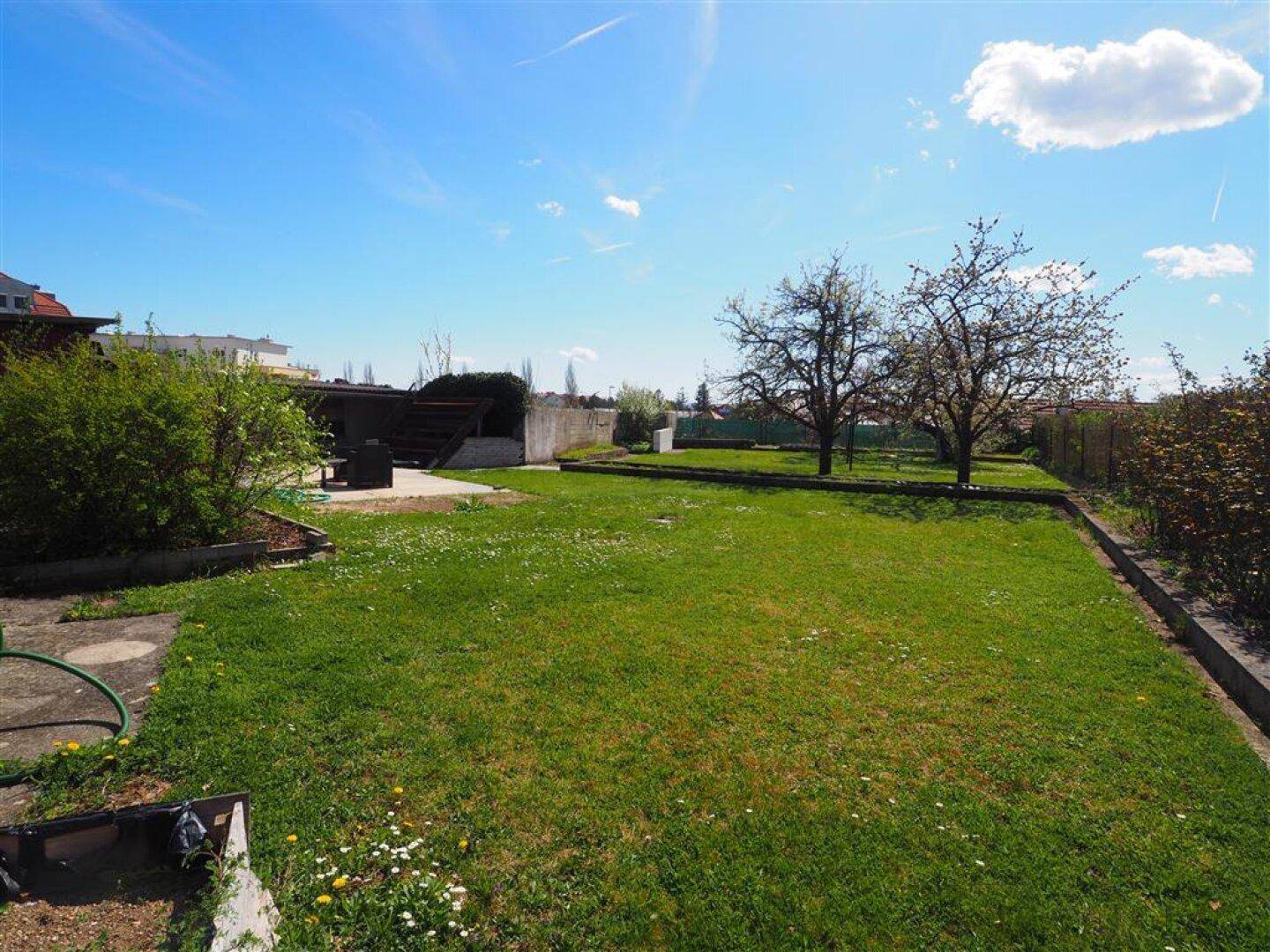 Weitläufiger Gartenbereich mit gepflegtem Rasen, blühenden Bäumen und einer kleinen Sitzgelegenheit im Hintergrund.