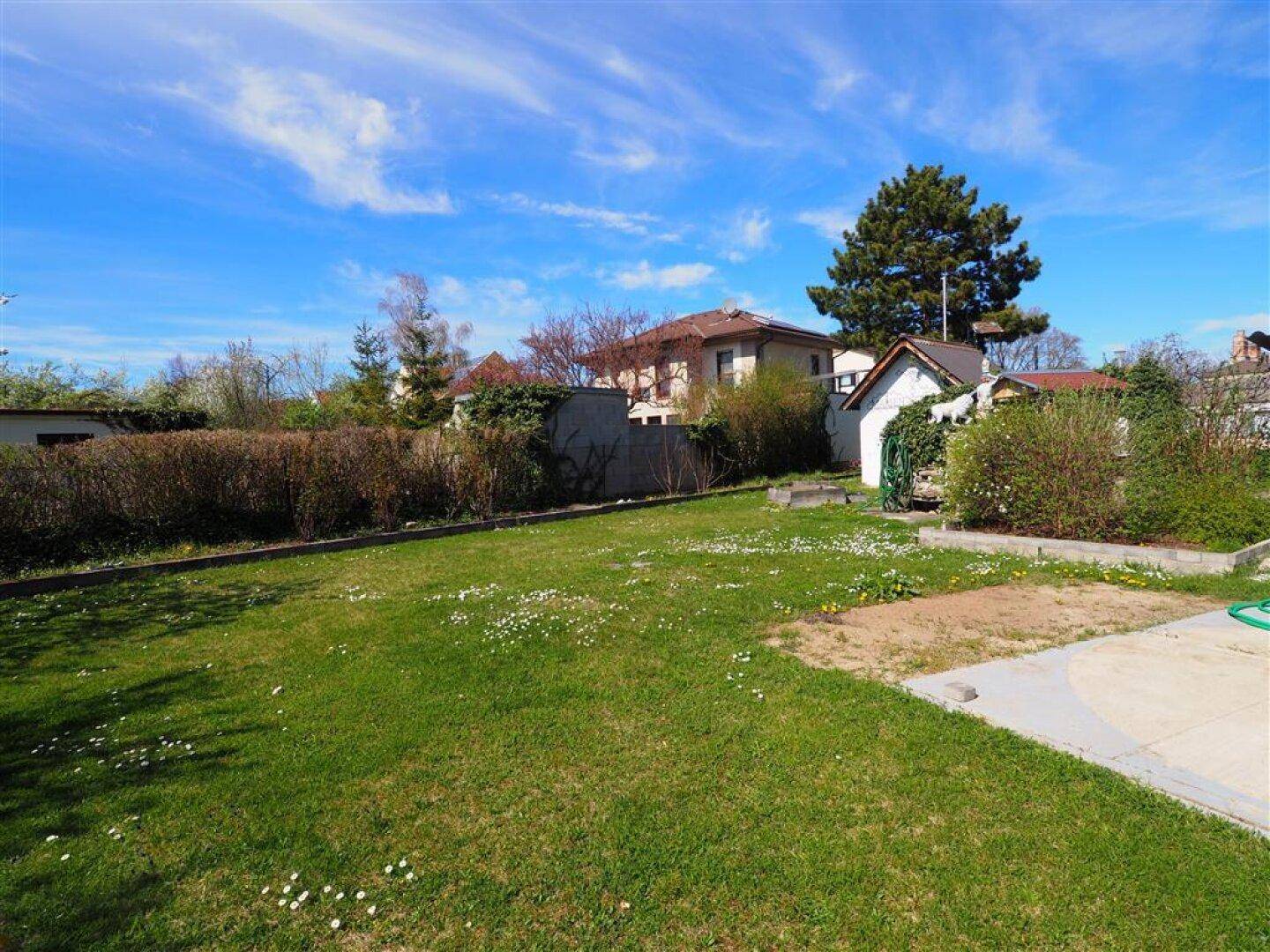 Gepflegter Garten mit Rasenfläche und Hecken, umgeben von benachbarten Häusern unter blauem Himmel.