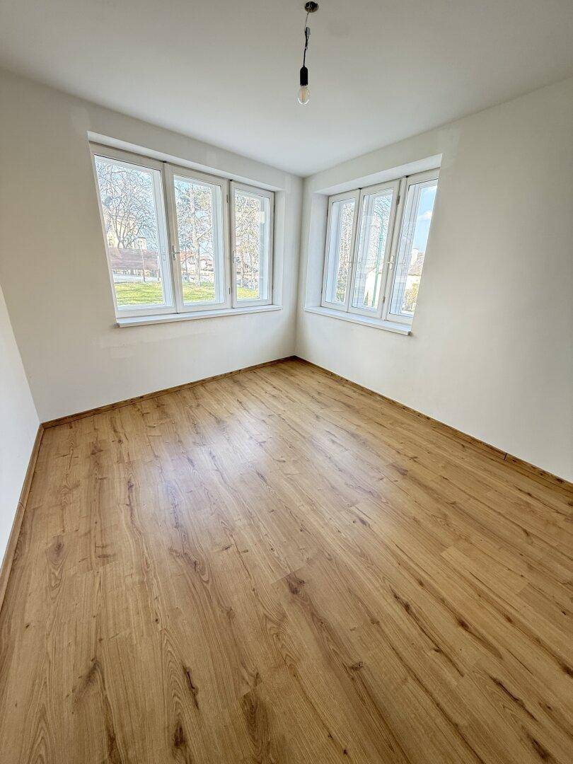 Helles Zimmer mit Holzoptik-Boden und zwei großen Fenstern.