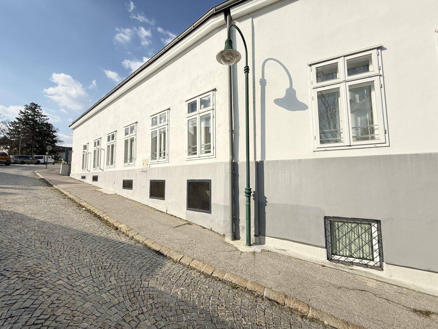 Lange weiße Fassade eines Gebäudes entlang einer gepflasterten Straße mit Laterne.