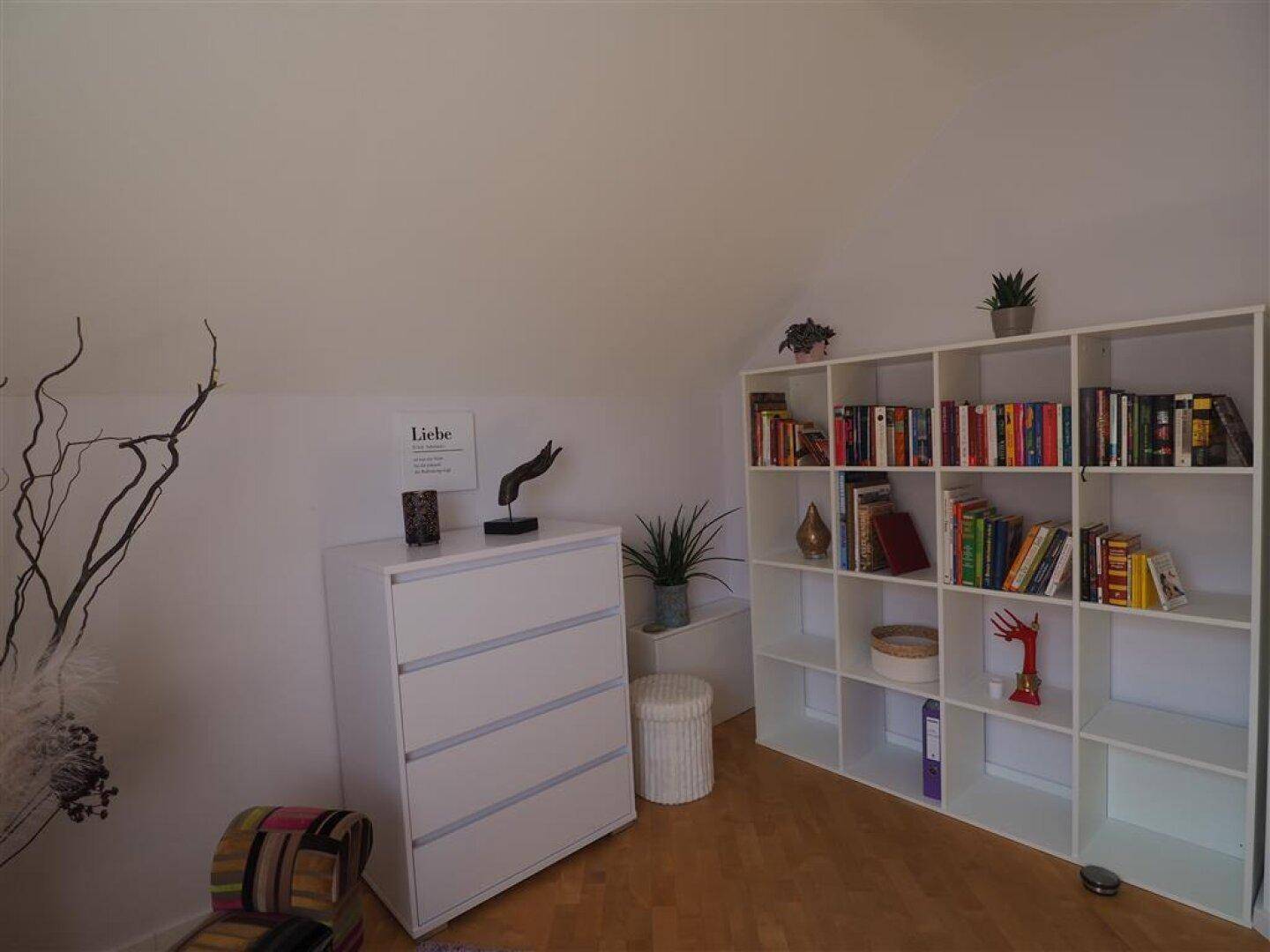 Helles Arbeitszimmer mit Bücherregalen, Kommode und Holzboden unter einer gemütlichen Dachschräge.