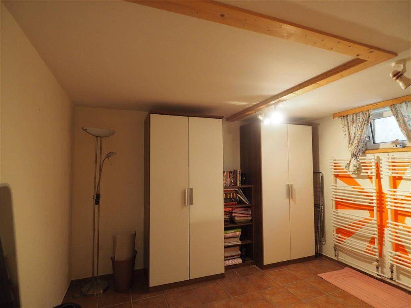 Kellerraum mit Kleiderschränken, Fenster und Heizkörper, ausgestattet mit Fliesenboden und Holzdeckenbalken.