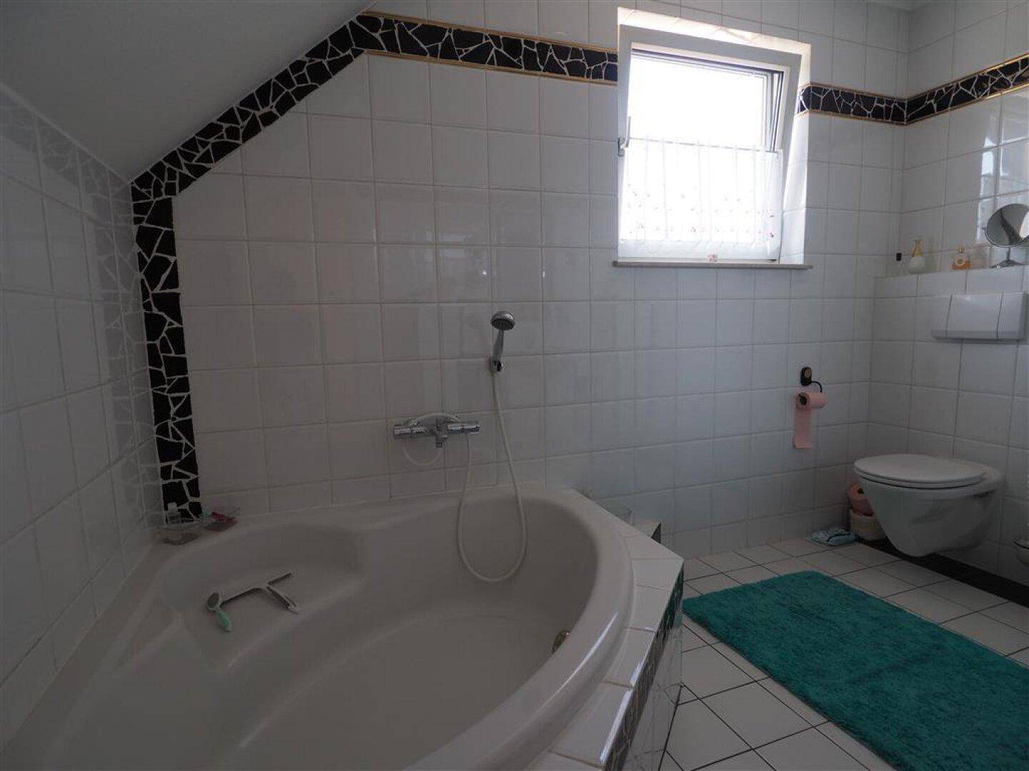 Badezimmer mit Eckbadewanne, Toilette und Fenster, gefliest in Weiß mit schwarzen Akzenten.