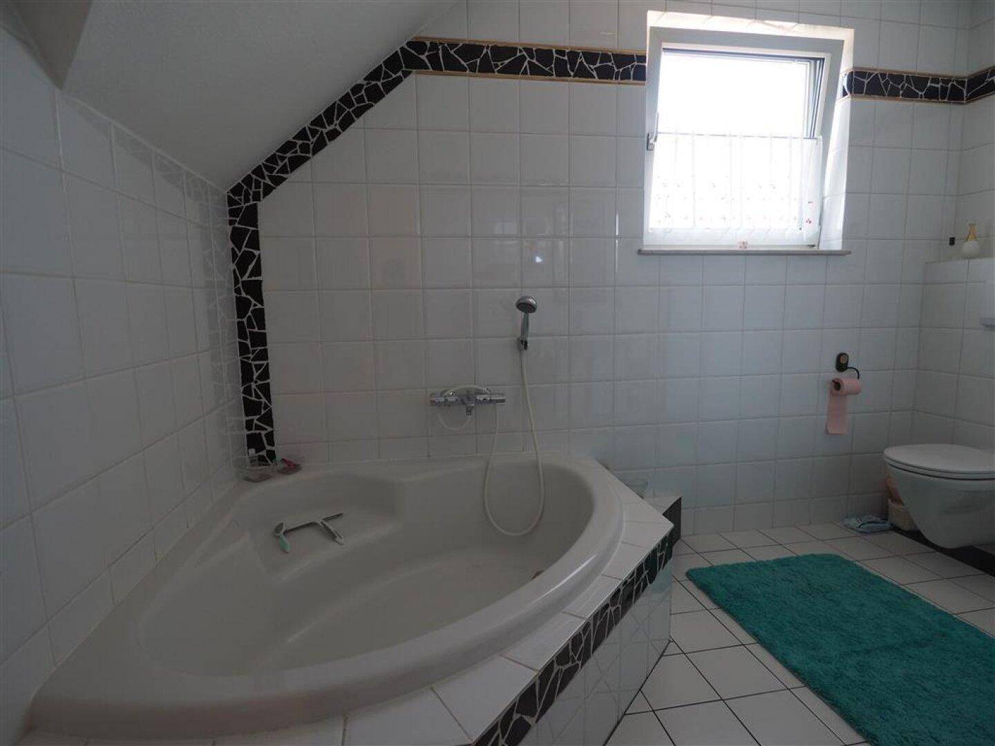 Helles Badezimmer mit Eckbadewanne, Toilette und Fenster, gefliest in Weiß mit schwarzen Akzenten.