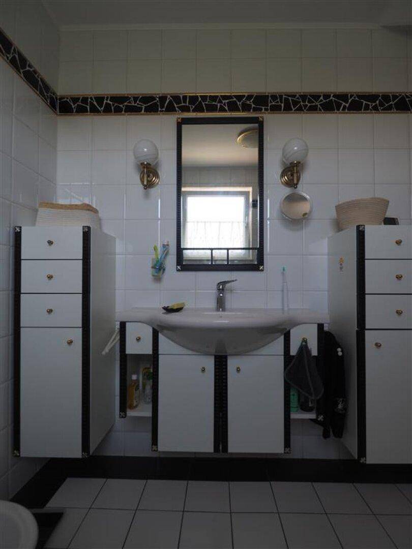 Badezimmer mit Waschbecken, Spiegel und weißen Schränken, gefliest in Weiß mit schwarzen Akzenten.