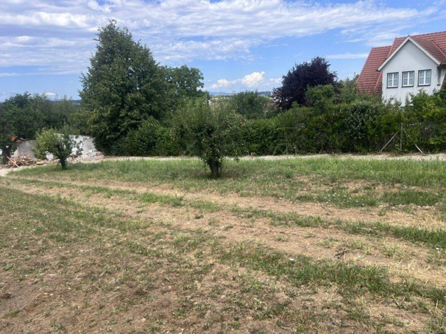 Offenes, grünes Grundstück mit leichter Hanglage und einem Haus im Hintergrund unter einem bewölkten Himmel.