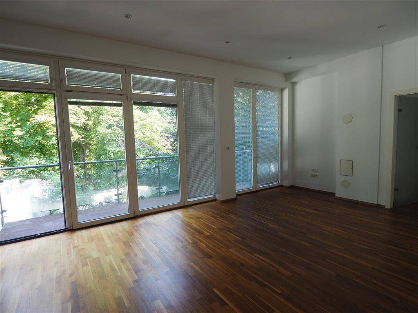 Heller Wohnbereich mit großen Fenstern, Holzboden und Zugang zum Balkon mit Grünblick.