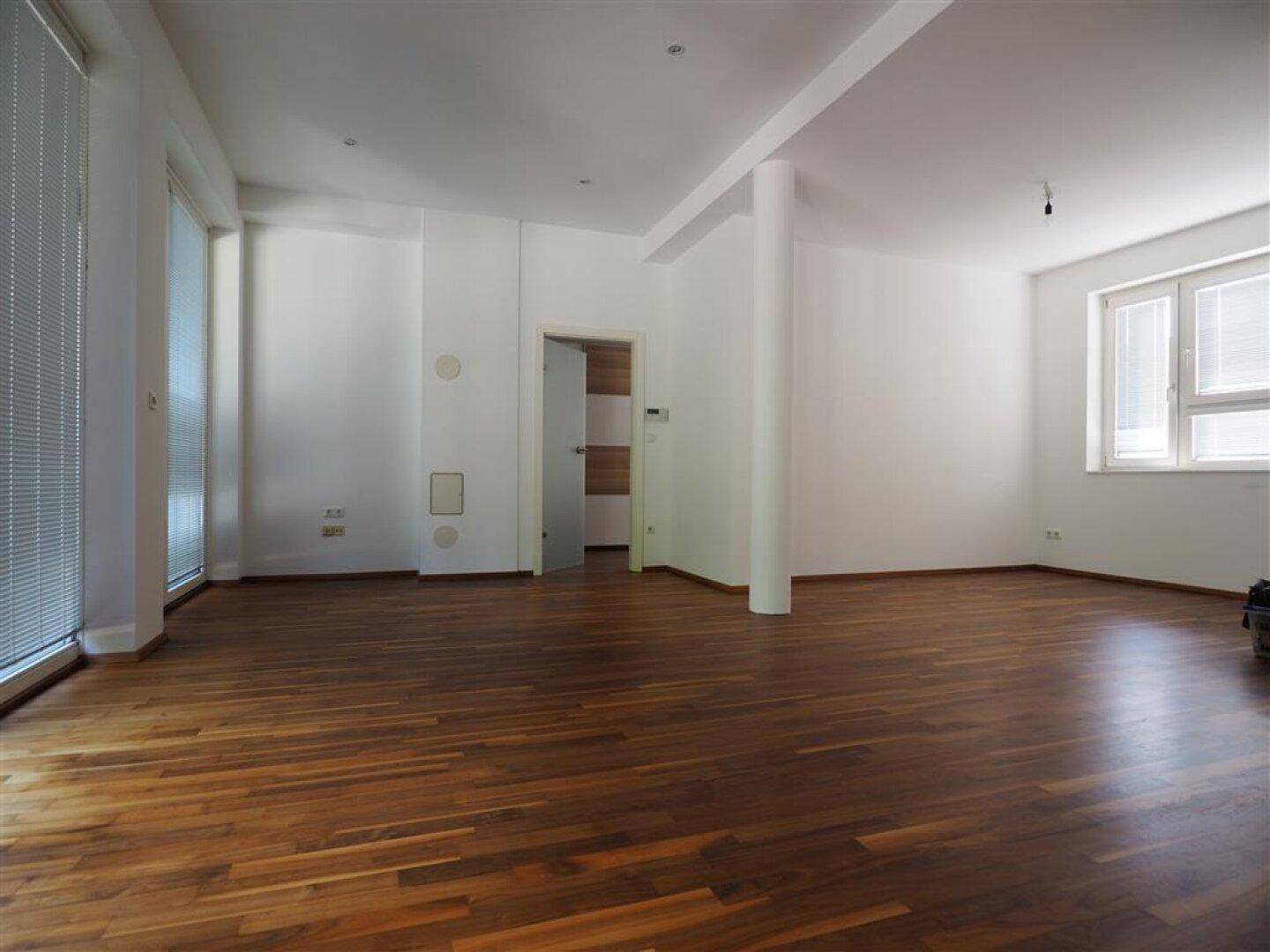 Heller Wohnbereich mit Holzboden, großen Fenstern und Zugang zu weiteren Räumen.