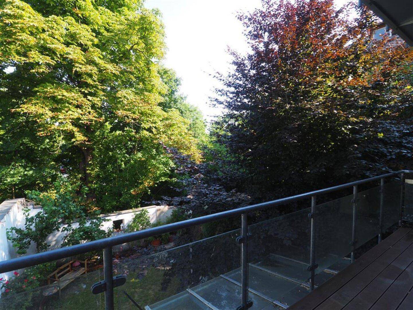 Balkon mit Glasgeländer und weitem Blick ins Grüne, umgeben von hohen Bäumen.
