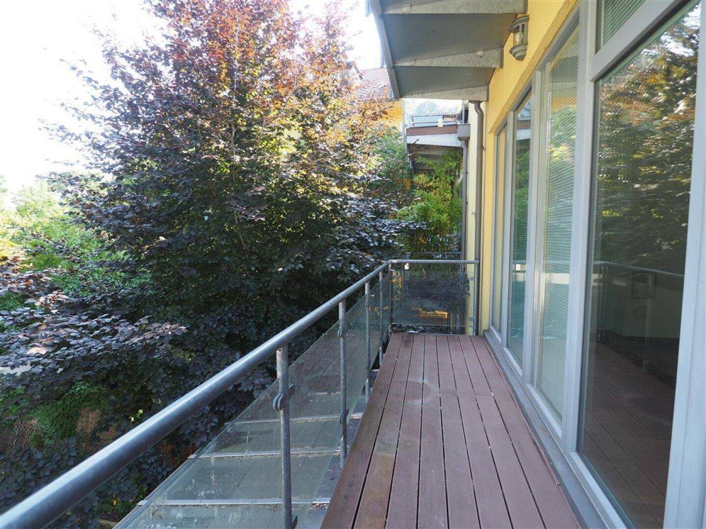 Langer Balkon mit Holzbelag und Glasgeländer, umgeben von üppigem Grün und Bäumen.