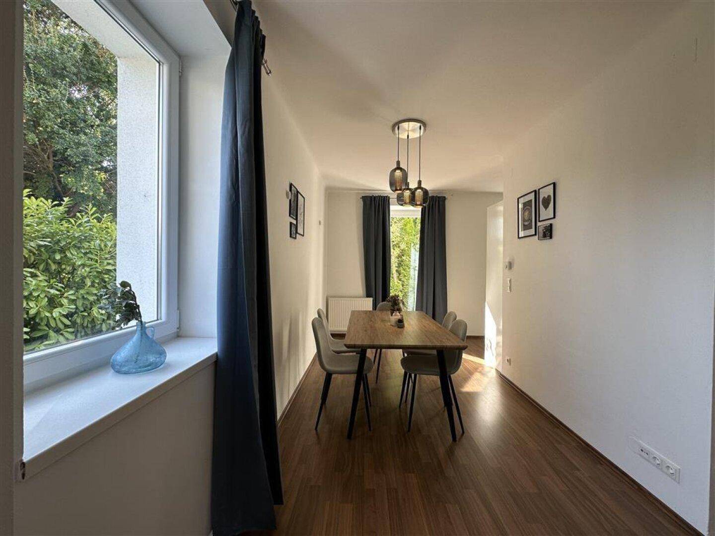 Langer Essbereich mit Holztisch und Stühlen, beleuchtet von einer modernen Hängelampe und Tageslicht.