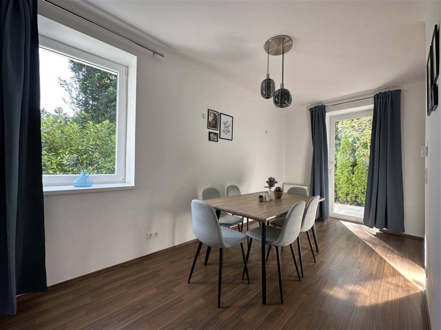 Heller Essbereich mit modernem Holztisch, grauen Stühlen und Blick ins Grüne durch große Fenster.
