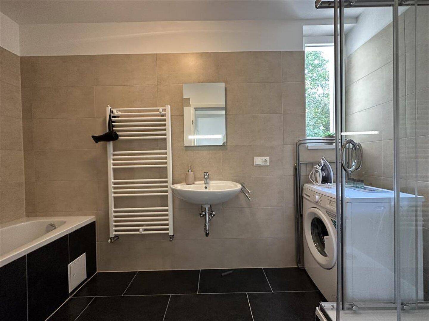 Helles Badezimmer mit Badewanne, Dusche, Waschbecken und Fenster, modern gefliest.