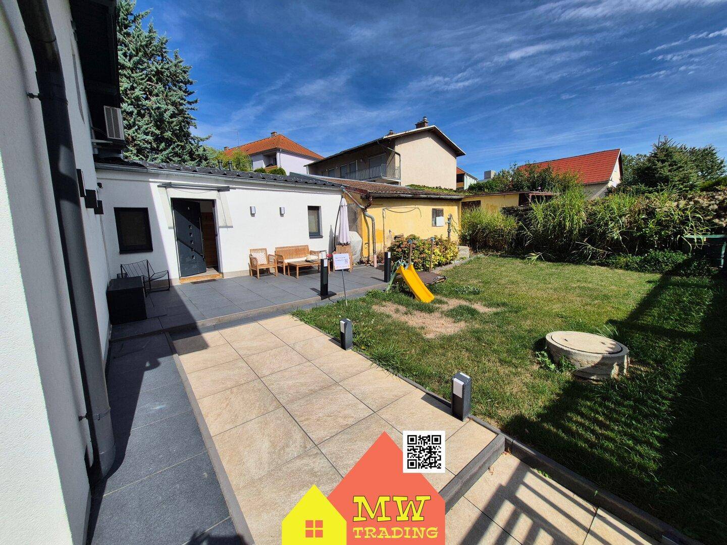 Weitläufiger Gartenbereich mit gepflasterter Terrasse, Rasen und Kinderspielgeräten.