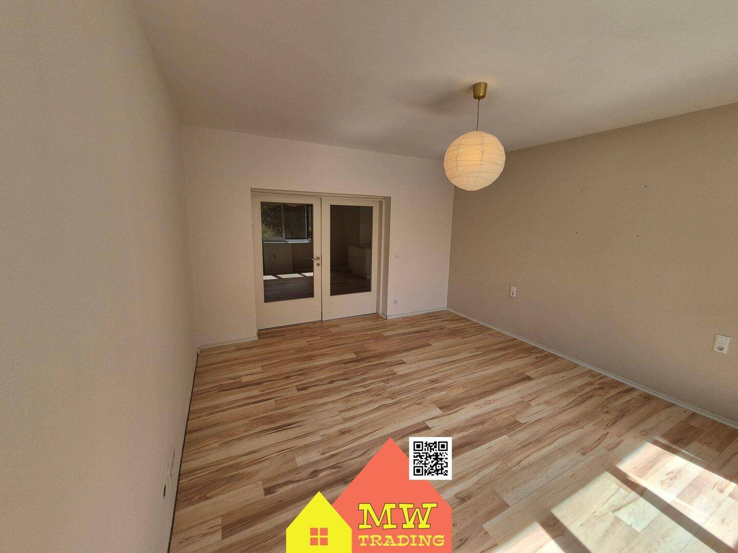 Helles Zimmer mit Holzboden und Zugang zu einem weiteren Raum durch Glastüren.