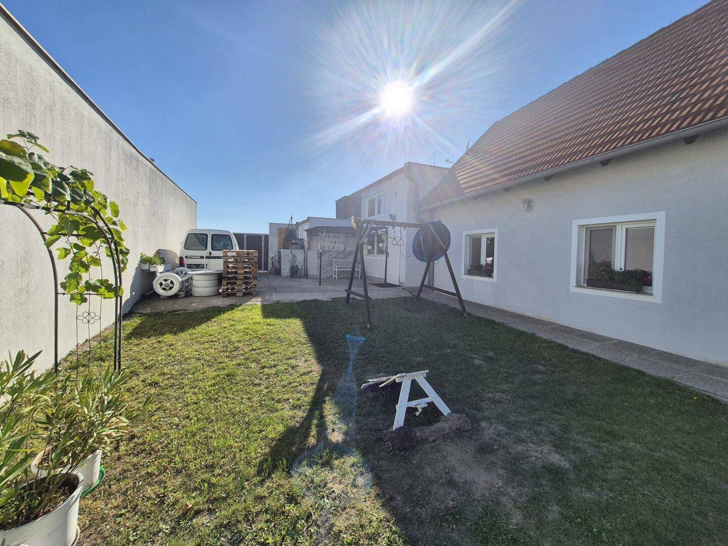 Gartenbereich neben dem Haus mit Rasenfläche und einigen Pflanzen, hell beleuchtet von der Sonne.