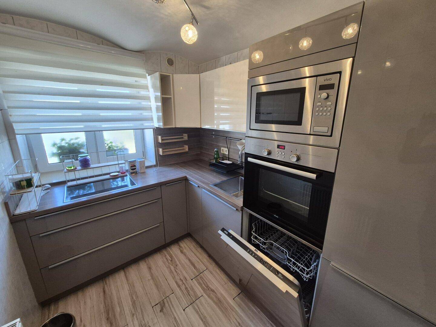 Helle Einbauküche mit modernen Geräten, geöffnetem Geschirrspüler und Fenster mit Jalousie.