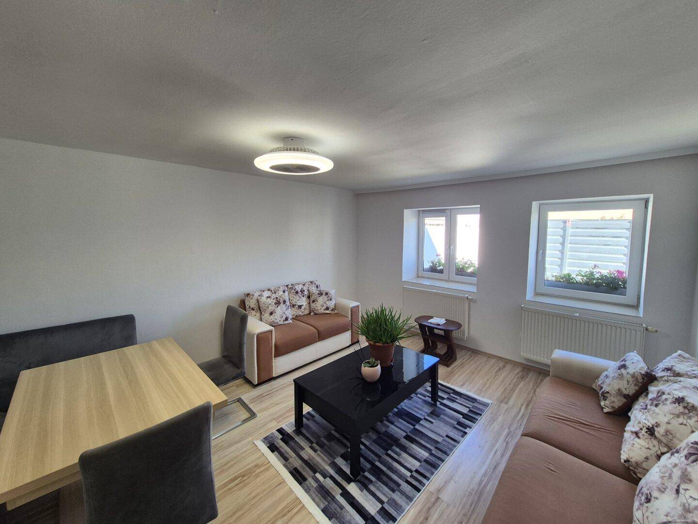 Heller Wohn- und Essbereich mit modernen Möbeln, Teppich und Fenstern mit Blick nach draußen.