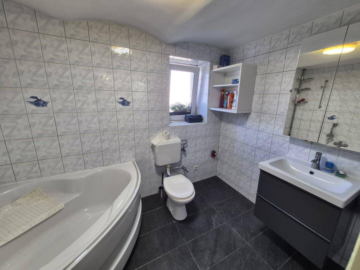 Badezimmer mit Eckbadewanne, Toilette, Waschtisch und hellen Fliesen an Wänden und Boden.