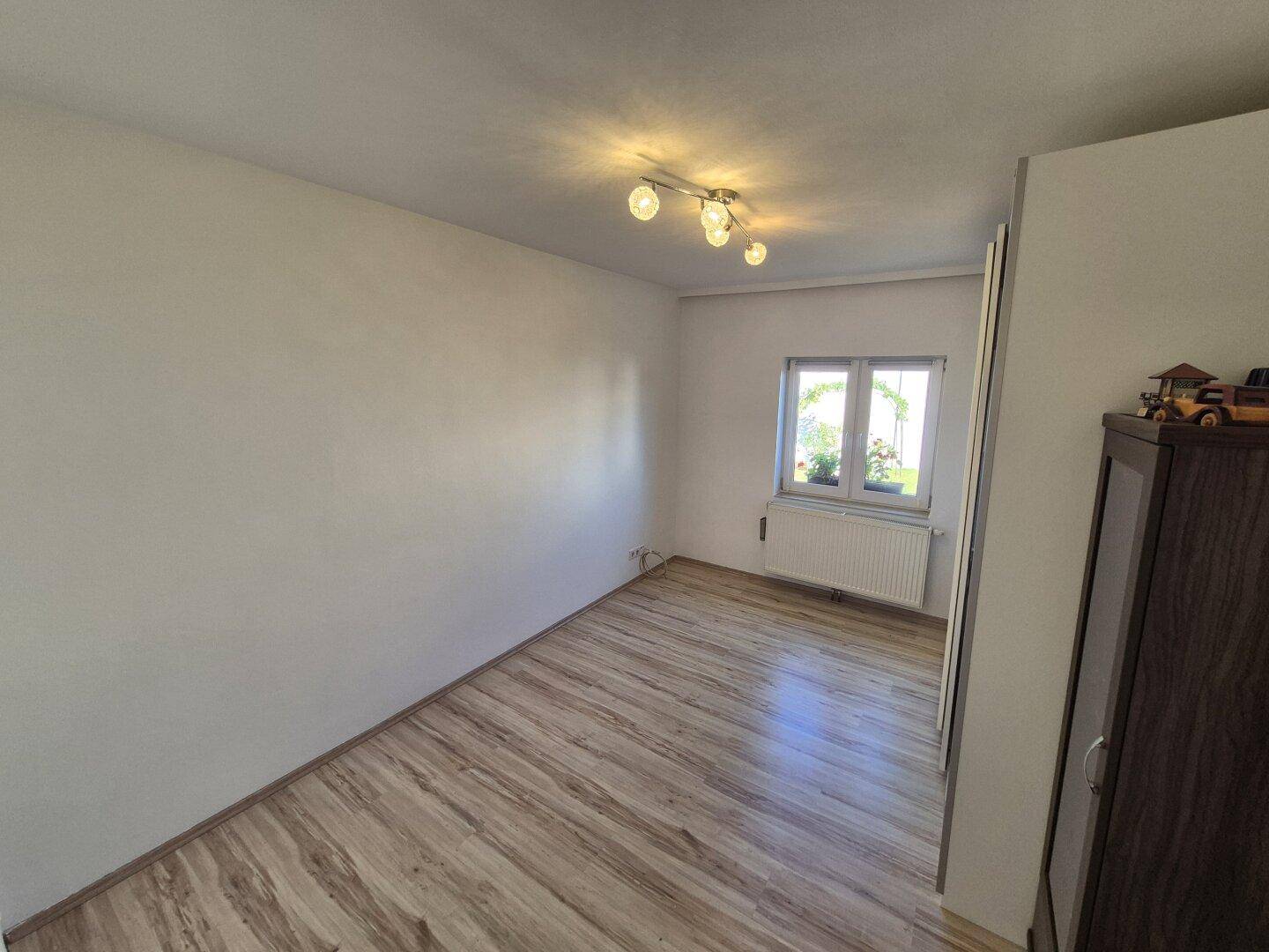 Helles, leeres Zimmer mit Holzoptik-Boden und einem Fenster, bereit zur individuellen Gestaltung.