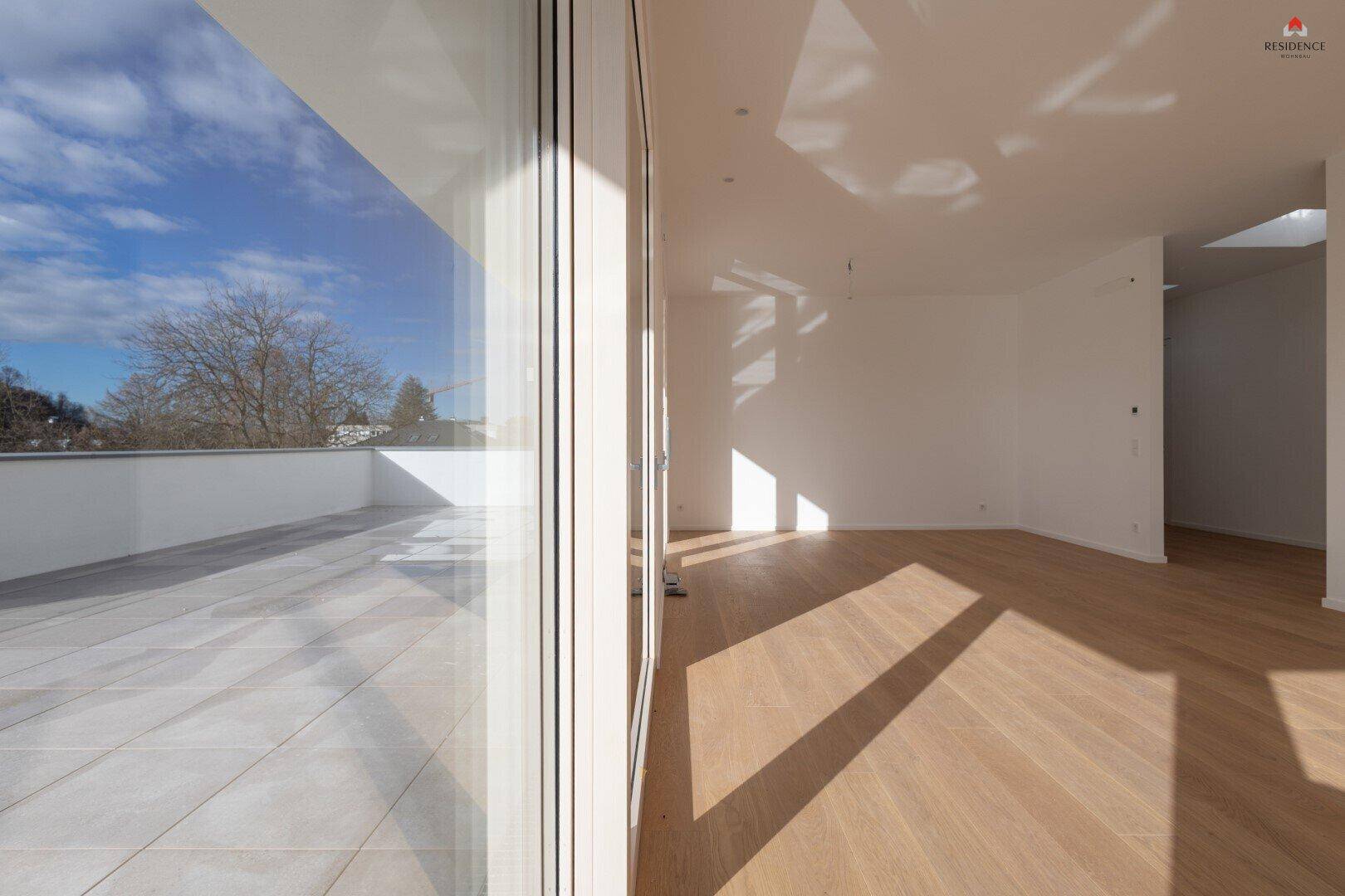 Heller Innenraum mit Holzboden und großem Fenster, das den Blick auf die geflieste Terrasse freigibt.