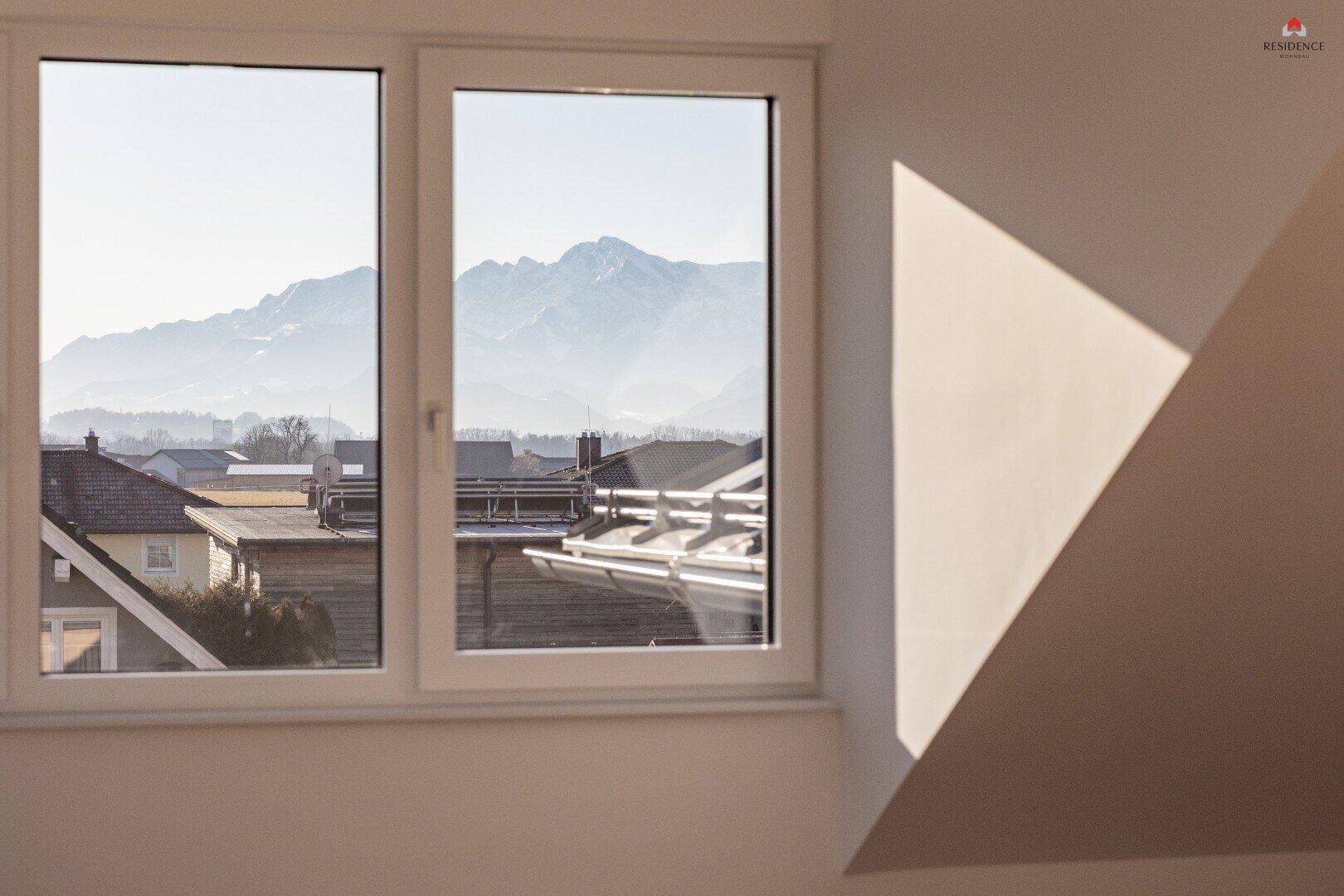 Fensterblick auf die Berglandschaft und umliegende Dächer von einem hellen Innenraum.