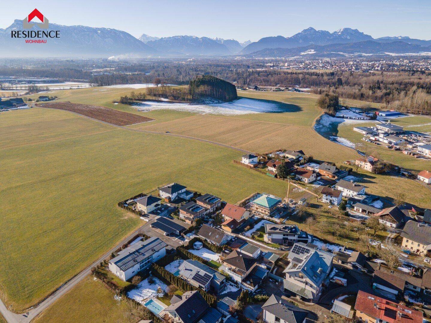 Luftaufnahme einer Wohnsiedlung mit Feldern und Bergen im Hintergrund.