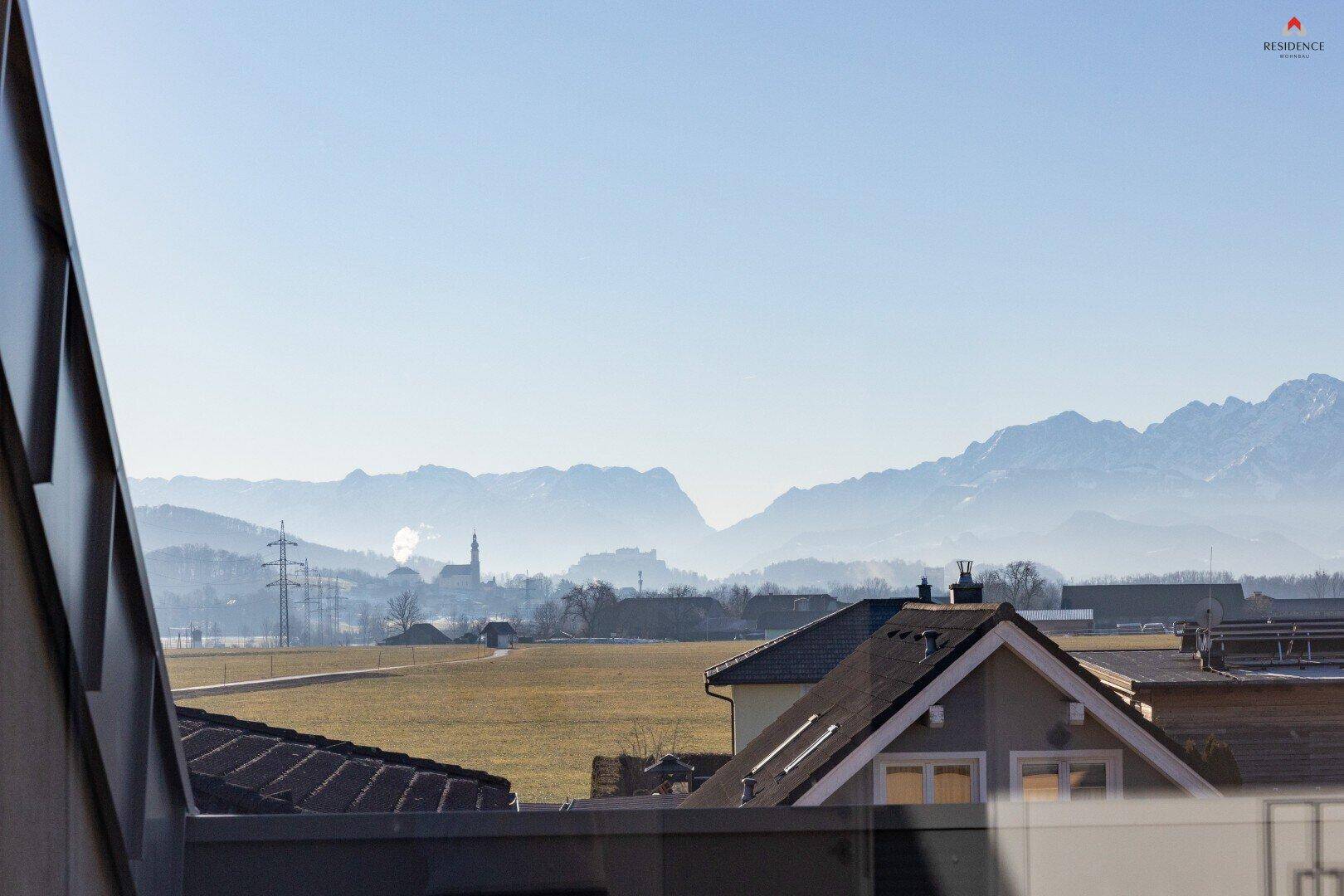 Weitläufiger Blick über Felder und Dächer auf die majestätische Berglandschaft in der Ferne.