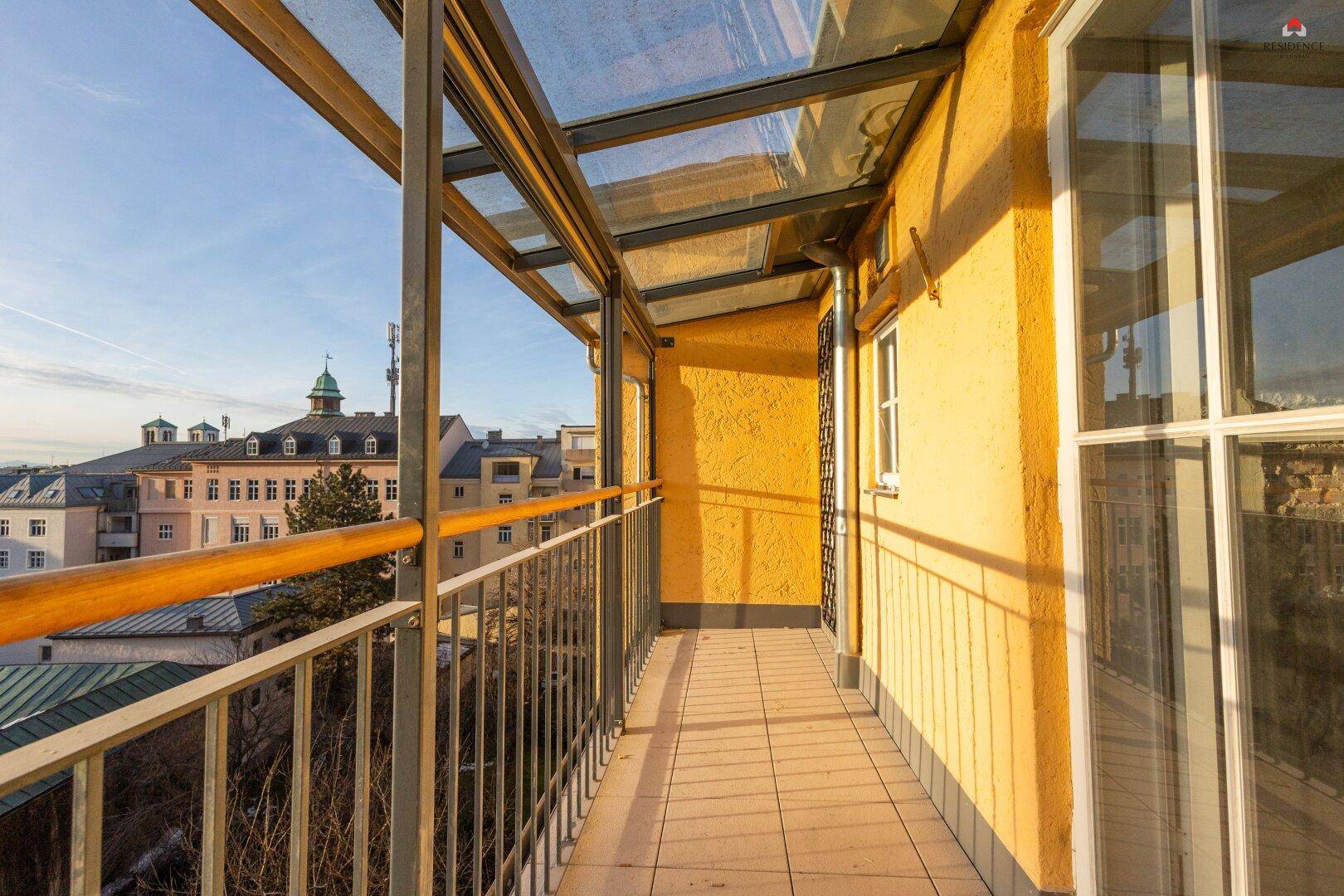 Sonniger, überdachter Balkon mit Fliesenboden und Blick auf die umliegenden Gebäude und Hügel.