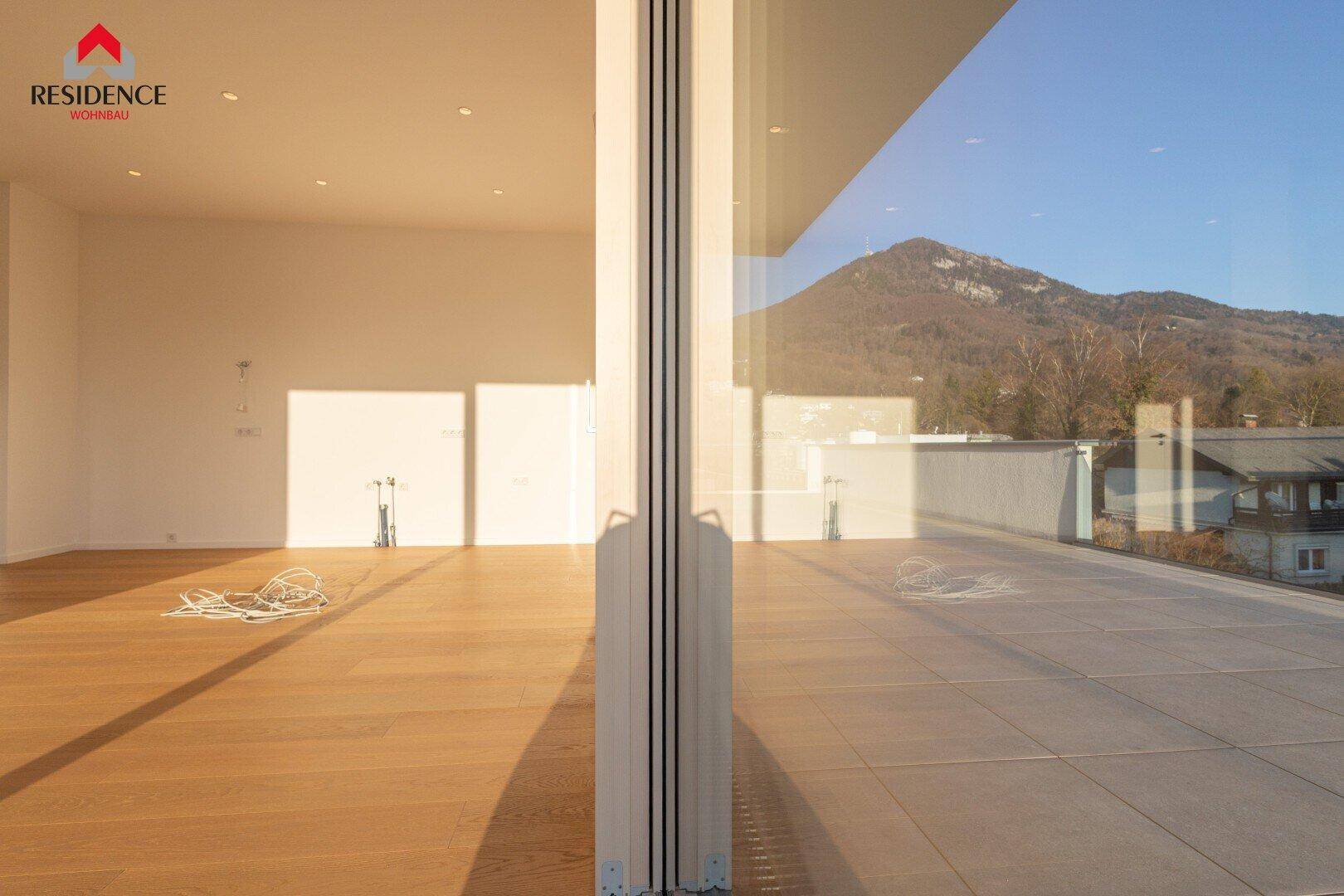 Heller Wohnraum mit Holzboden und großzügigen Glasfronten, die den Blick nach draußen öffnen.