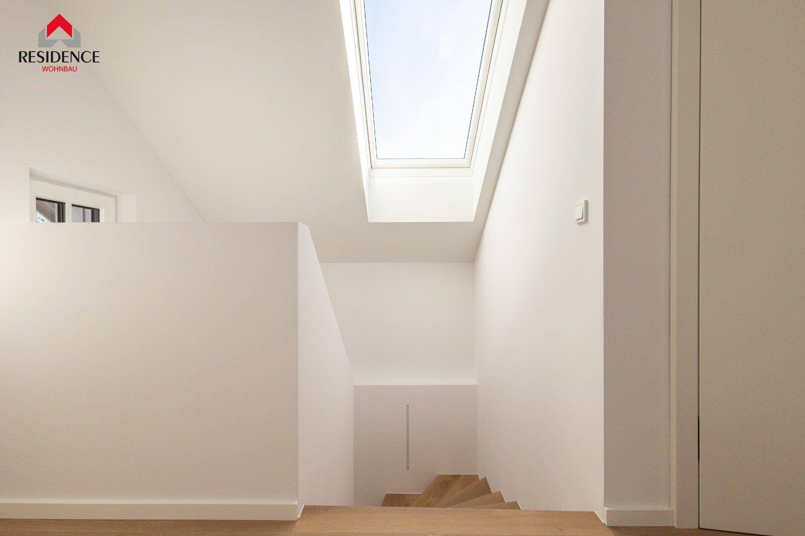 Heller Treppenaufgang mit Holzboden, weiß gestrichenen Wänden und einem großen Dachfenster.