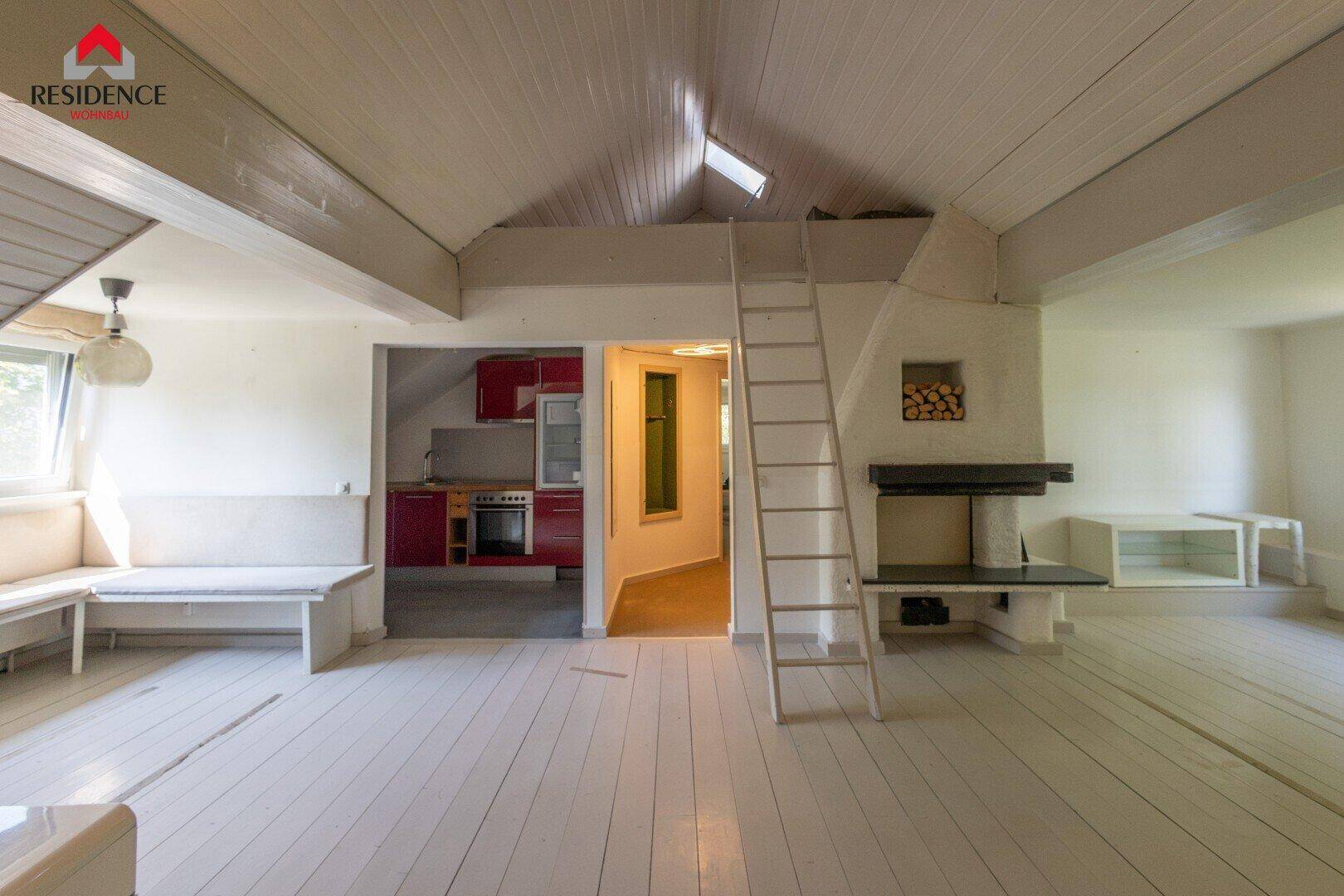 Heller Wohnbereich mit Kamin, Holzfußboden und Zugang zu einer roten Küche sowie einer Leiter zum Dachboden.