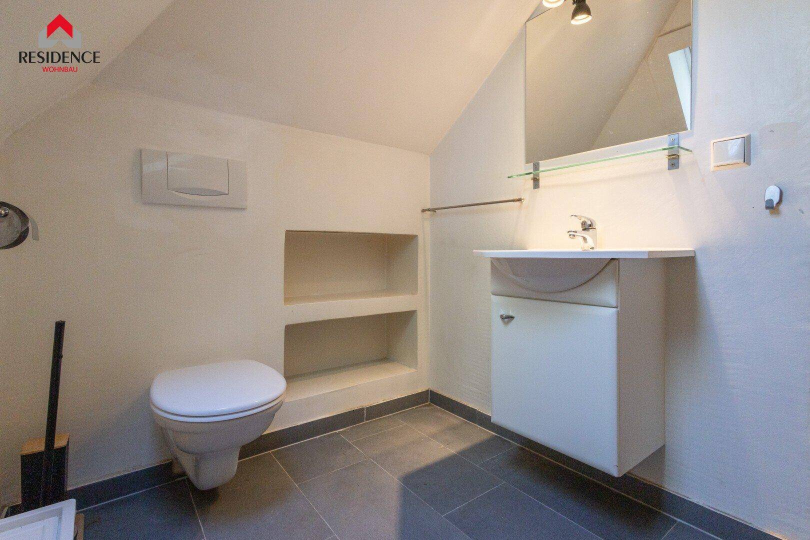 Modernes Badezimmer mit wandhängendem WC, Einzelwaschbecken und dunklen Fliesen, unter einer Dachschräge gelegen.