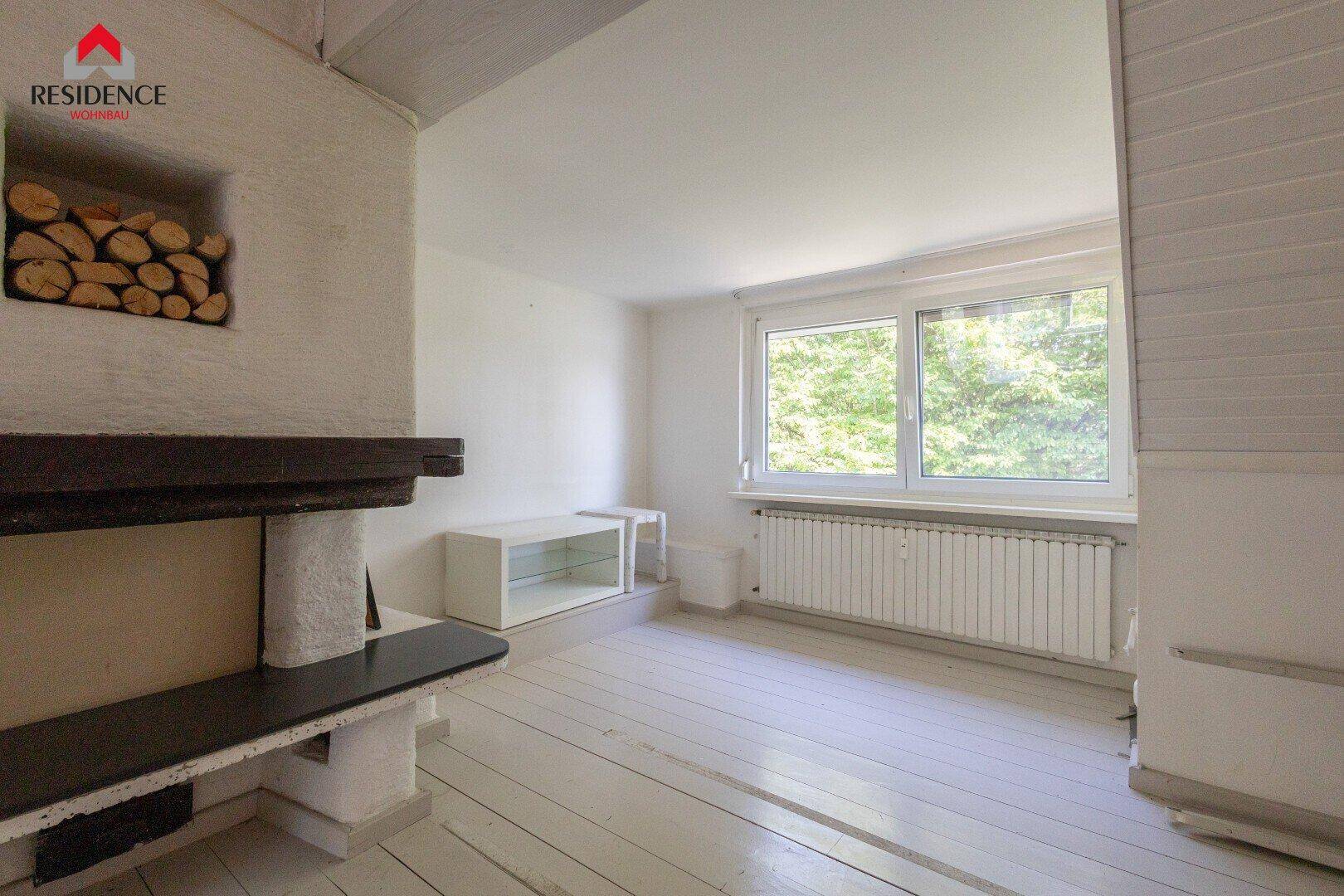 Detailansicht des Kamins mit Holzscheiten und eines Fensters mit Heizkörper, auf weißem Holzfußboden.