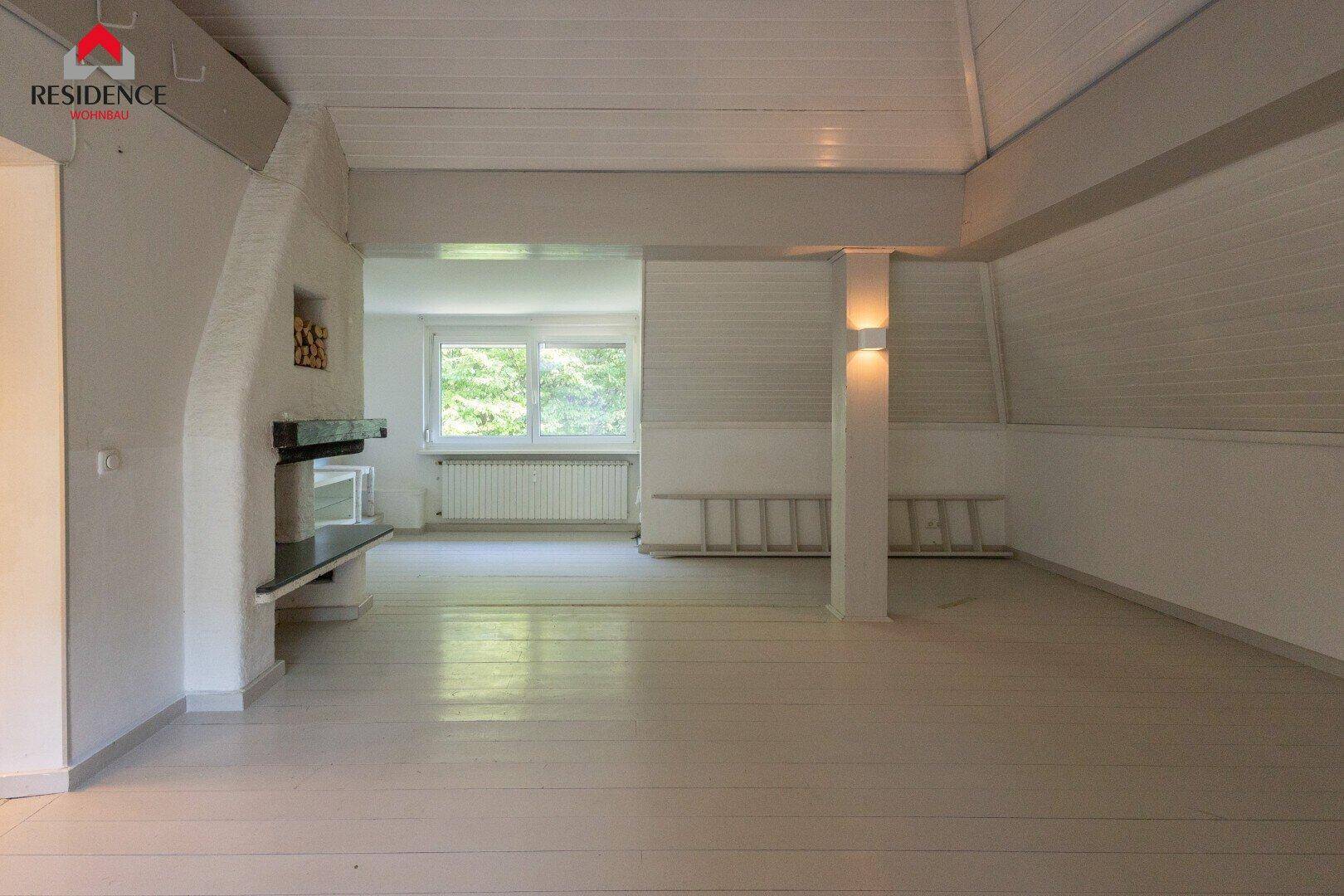 Geräumiger Wohnbereich mit Kamin, weißem Holzfußboden und großen Fenstern, die viel Licht hereinlassen.