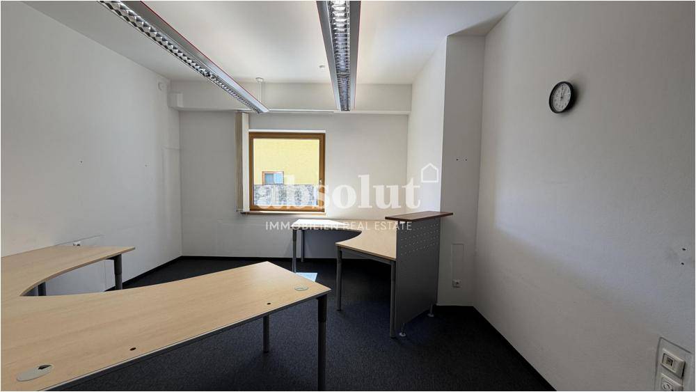 Helles Büro mit mehreren Schreibtischen, Teppichboden und einer Wanduhr.