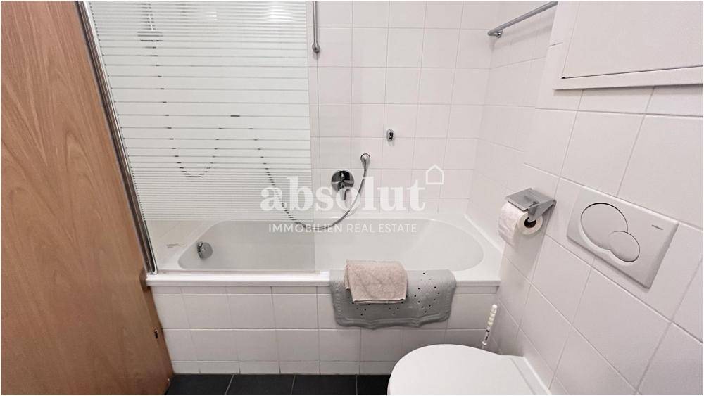 Helles Badezimmer mit Badewanne, Duschwand und weißen Fliesen, funktional eingerichtet.