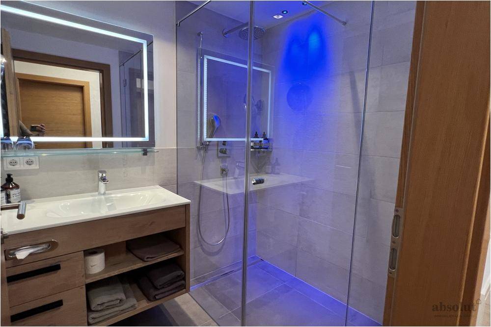 Elegantes Badezimmer mit großer Glasdusche, beleuchtetem Spiegel und Holzwaschtisch.